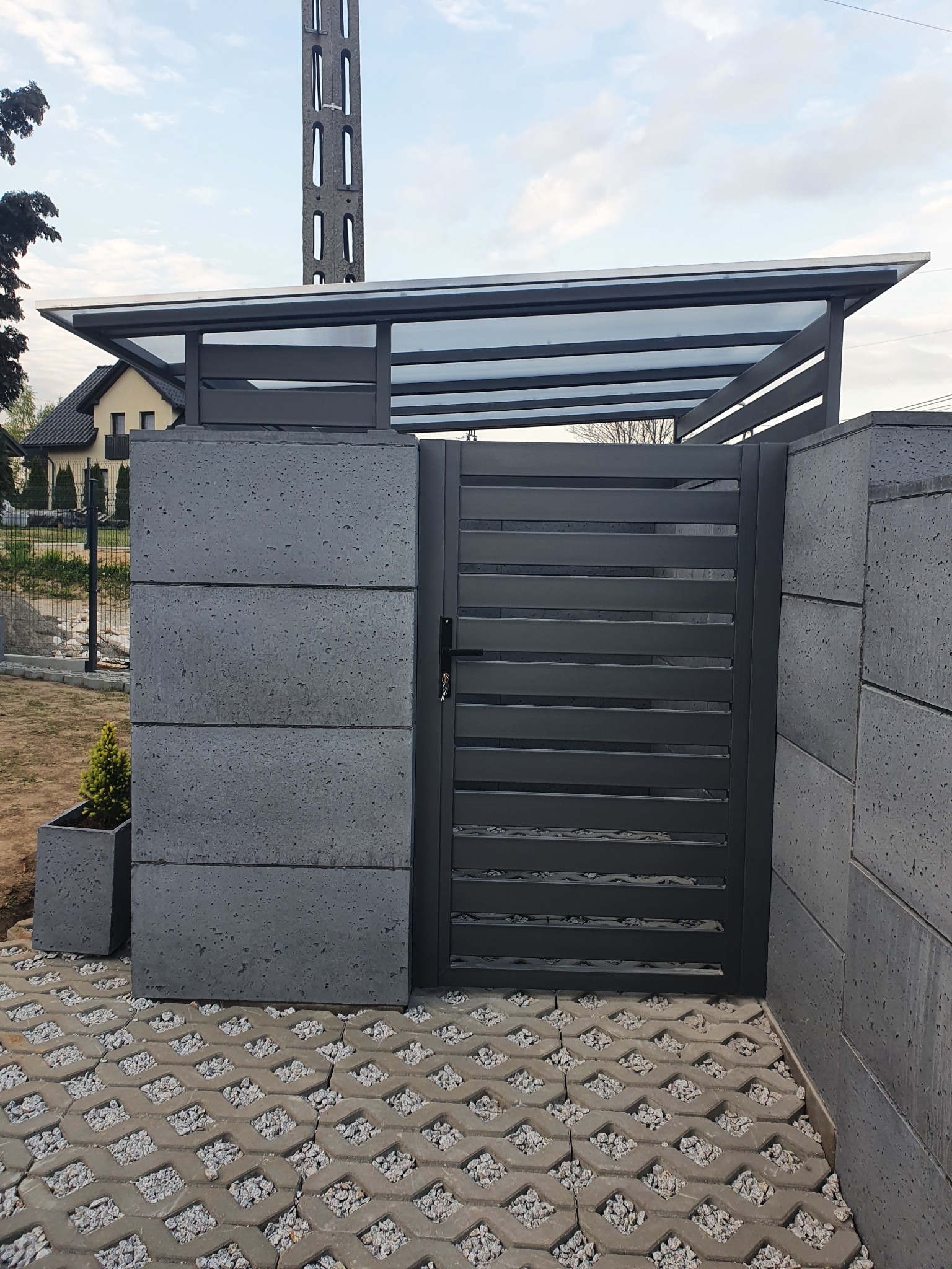 Nowoczesna, pozioma brama wjazdowa w kolorze grafitowym z zadaszeniem z przezroczystego tworzywa, osadzona na betonowych słupkach i kostce ażurowej wypełnionej kamieniami. Obok donica z małą...