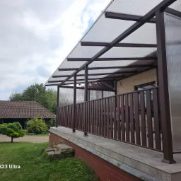 Zadaszony taras z brązowymi, metalowymi poręczami i przezroczystym zadaszeniem z poliwęglanu, widok z zewnątrz na zadbany ogród z domem w tle.