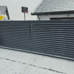 Nowoczesna, szara brama wjazdowa lamelowa z furtką i skrzynką na listy, widoczna na tle ośnieżonego podjazdu i zabudowań.