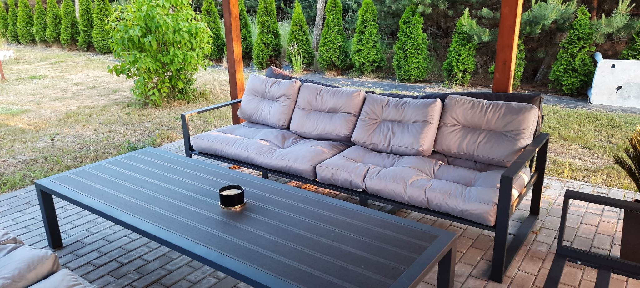 Zestaw mebli ogrodowych z metalowym stelażem i szarymi poduchami, stół z czarnym świecznikiem, na tle tuj i trawnika.