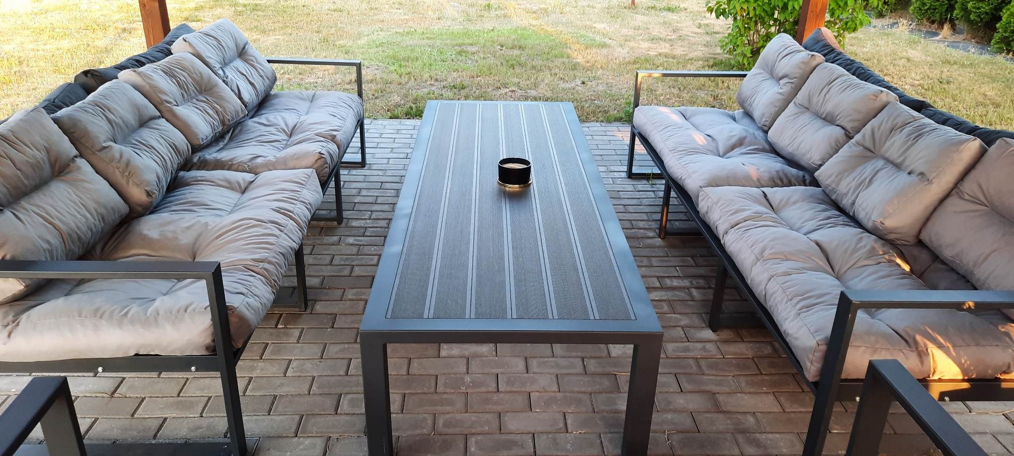 Zestaw mebli ogrodowych z metalową ramą i miękkimi poduchami w kolorze szarym, stół z blatem o fakturze drewna, na brukowanym tarasie.