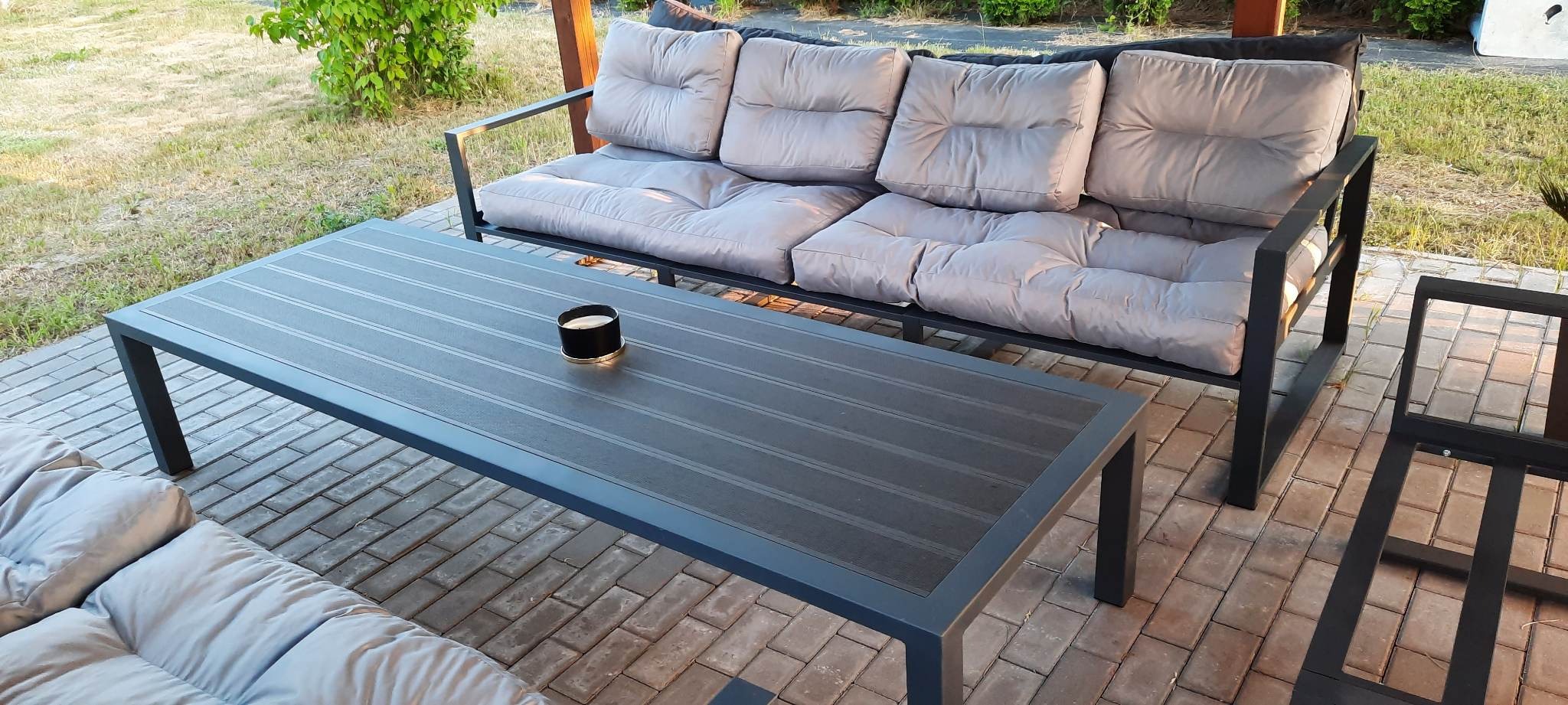 Zestaw mebli ogrodowych z metalową ramą i szarymi poduszkami na tarasie z kostki brukowej, z czarną świecą na stole.