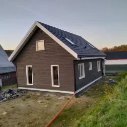 Nowoczesny dom z ciemną, drewnianą elewacją i czarnym dachem z oknami dachowymi, widok z zewnątrz na tle zieleni i innych budynków.