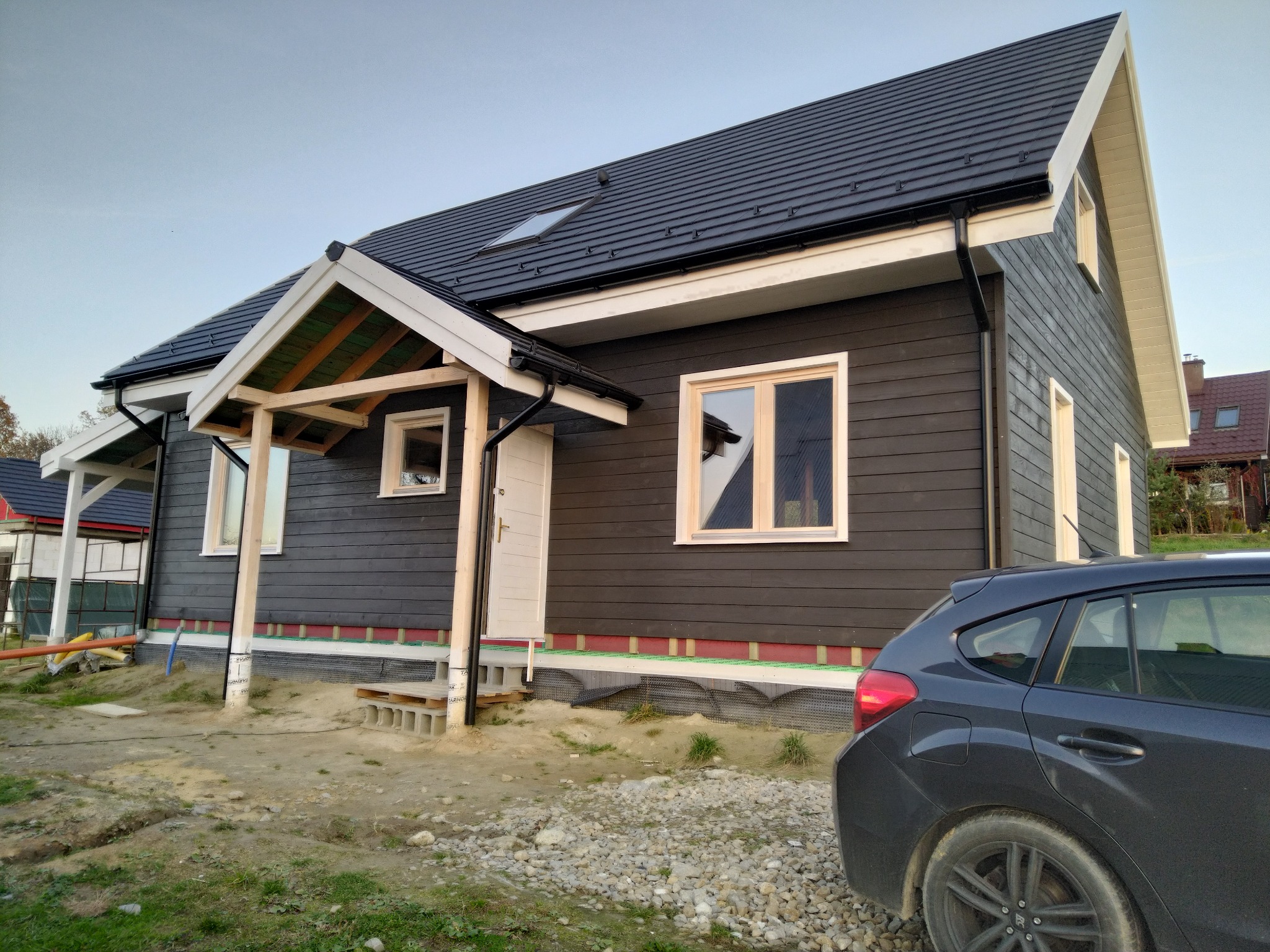 Niedokończony dom szkieletowy z ciemną elewacją i białymi obramowaniami okien, widoczne fundamenty i fragment samochodu na pierwszym planie.