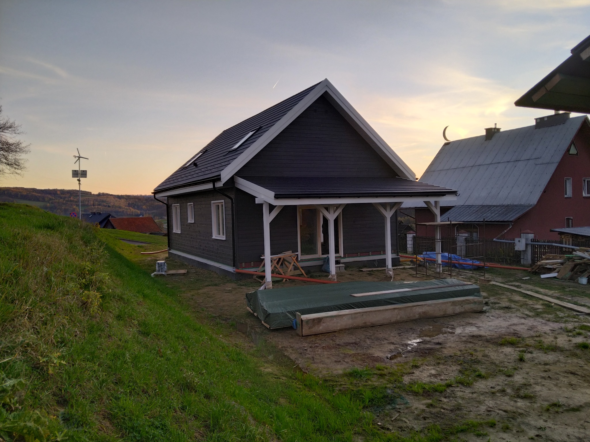 Nowoczesny, ciemnoszary dom z białymi obramowaniami okien i zadaszonym gankiem w trakcie budowy, widoczny w krajobrazie z turbiną wiatrową w tle.