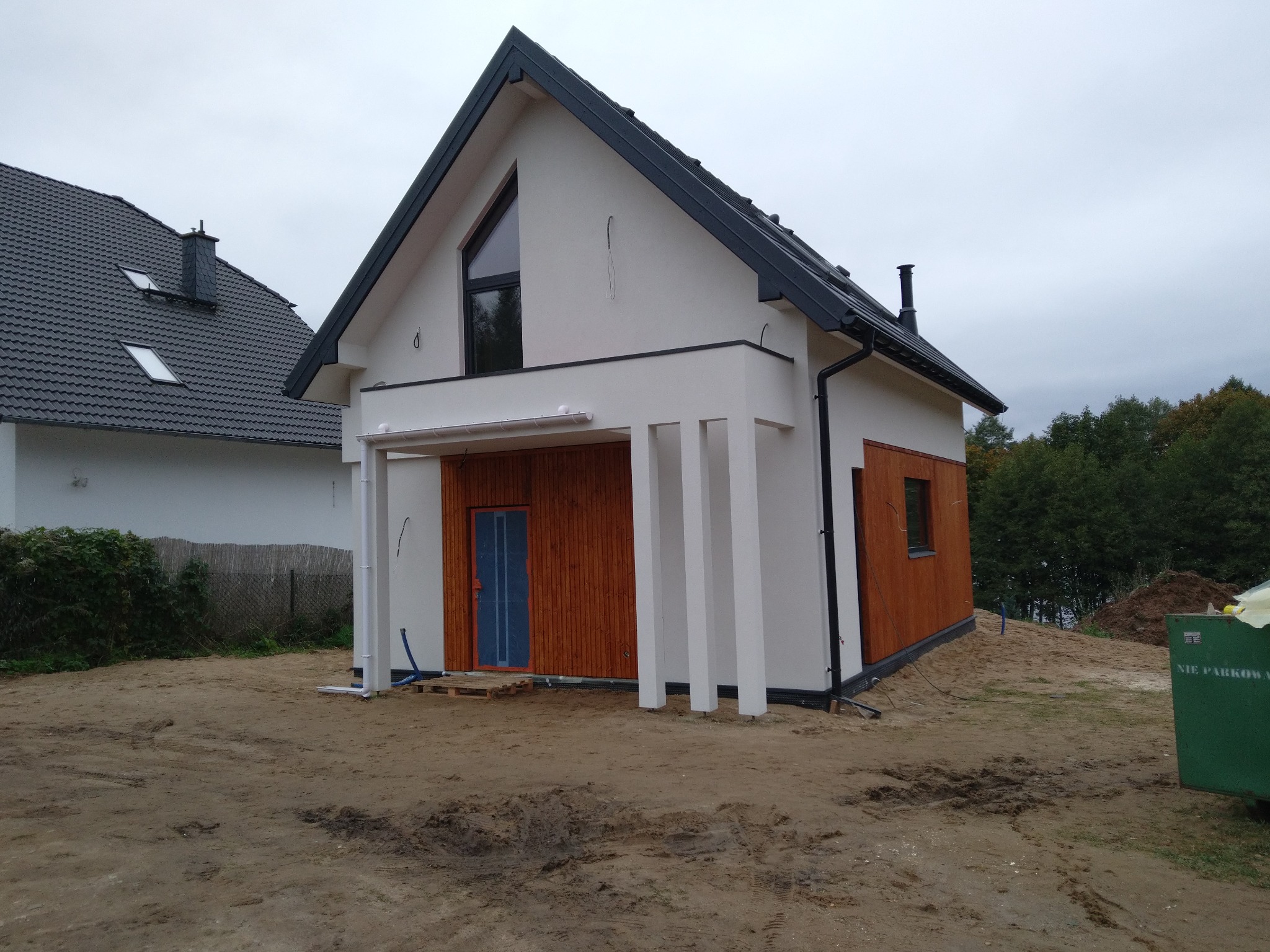 Nowoczesny dom z białym tynkiem i drewnianą elewacją, z widoczną stolarką okienną w kolorze czarnym, drzwi wejściowe zabezpieczone folią, teren wokół domu w trakcie prac ziemnych.