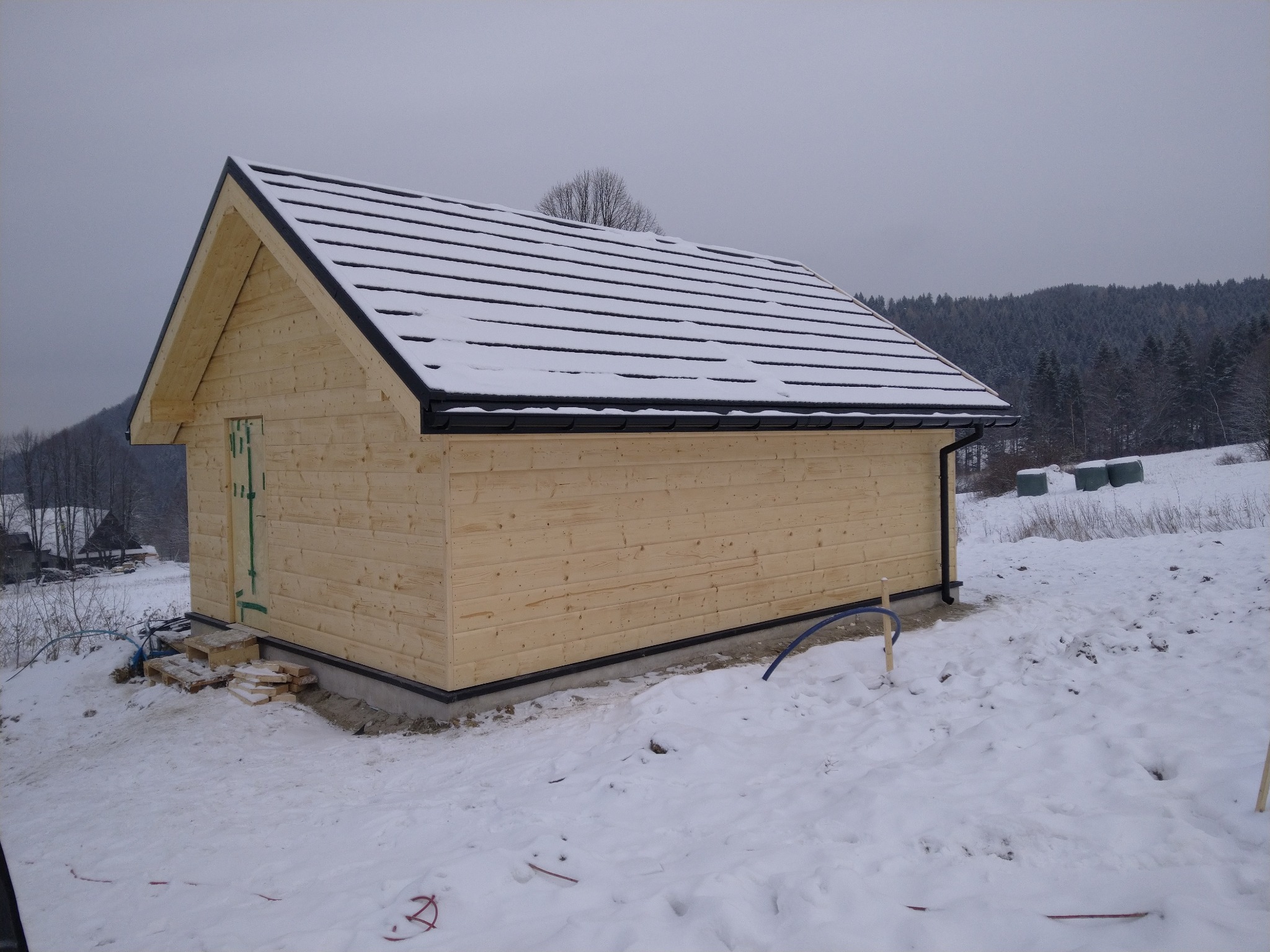 Surowy drewniany budynek z dwuspadowym dachem pokrytym śniegiem, widoczne tymczasowe schody z palet i rury na tle zasypanego śniegiem terenu i lasu.