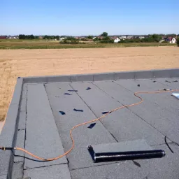 Nowa papa termozgrzewalna na płaskim dachu, częściowo zabezpieczona łatami, z widocznym palnikiem i przewodem gazowym, w tle pole i zabudowania.