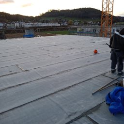 Dr.dachwalkowiak - Widok z góry na płaski dach pokryty papą termozgrzewalną, z widocznym pracownikiem w oddali, pomarańczowym kaskiem i żółtym dźwigiem w tle, podczas montażu na budowie.