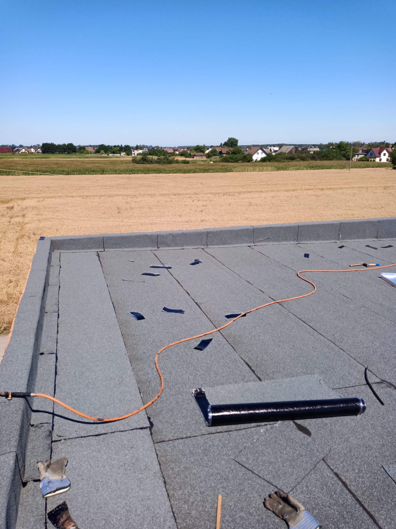 Nowa papa termozgrzewalna na płaskim dachu, częściowo zabezpieczona łatami, z widocznym palnikiem i przewodem gazowym, w tle pole i zabudowania.