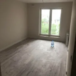 Pusty pokój po remoncie: świeżo ułożone panele podłogowe z efektem drewna, jasne ściany, widok na zieleń za oknem, butelka wody na podłodze.