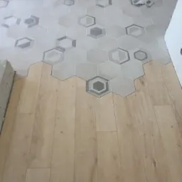 Połączenie podłogi z płytek heksagonalnych w odcieniach szarości z panelami podłogowymi o jasnym, naturalnym odcieniu drewna, widoczne niedokończone krawędzie i fragmenty betonu.