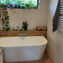 Nowoczesna łazienka z wolnostojącą wanną, drewnopodobną półką z roślinami i grzejnikiem drabinkowym z szarym ręcznikiem.