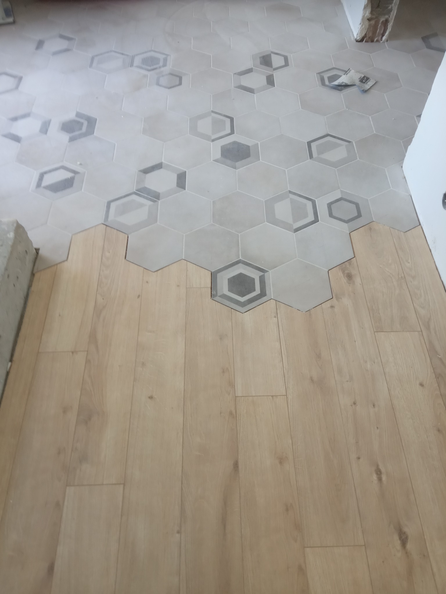 Połączenie podłogi z płytek heksagonalnych w odcieniach szarości z panelami podłogowymi o jasnym, naturalnym odcieniu drewna, widoczne niedokończone krawędzie i fragmenty betonu.