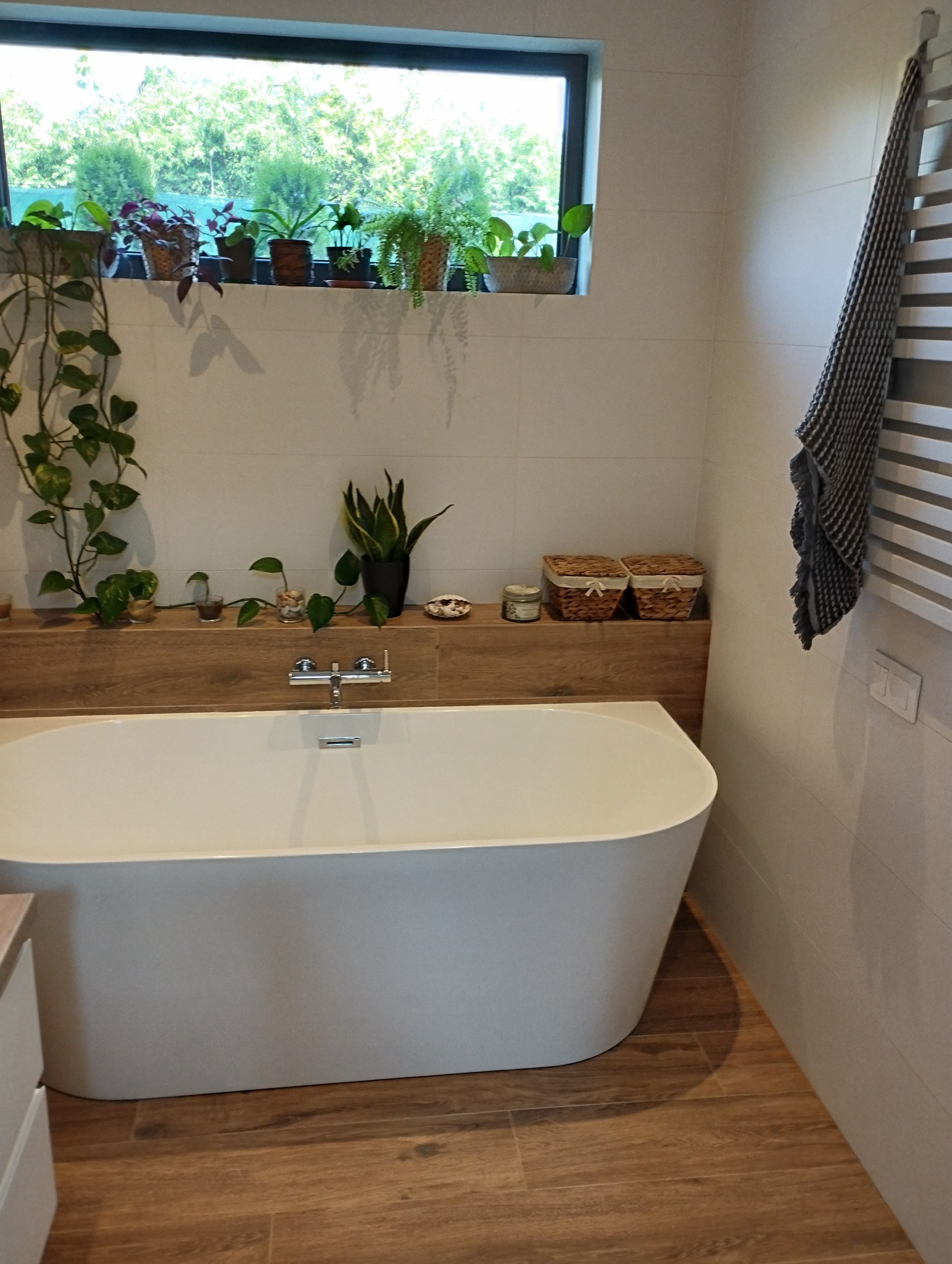 Nowoczesna łazienka z wolnostojącą wanną, drewnopodobną półką z roślinami i grzejnikiem drabinkowym z szarym ręcznikiem.