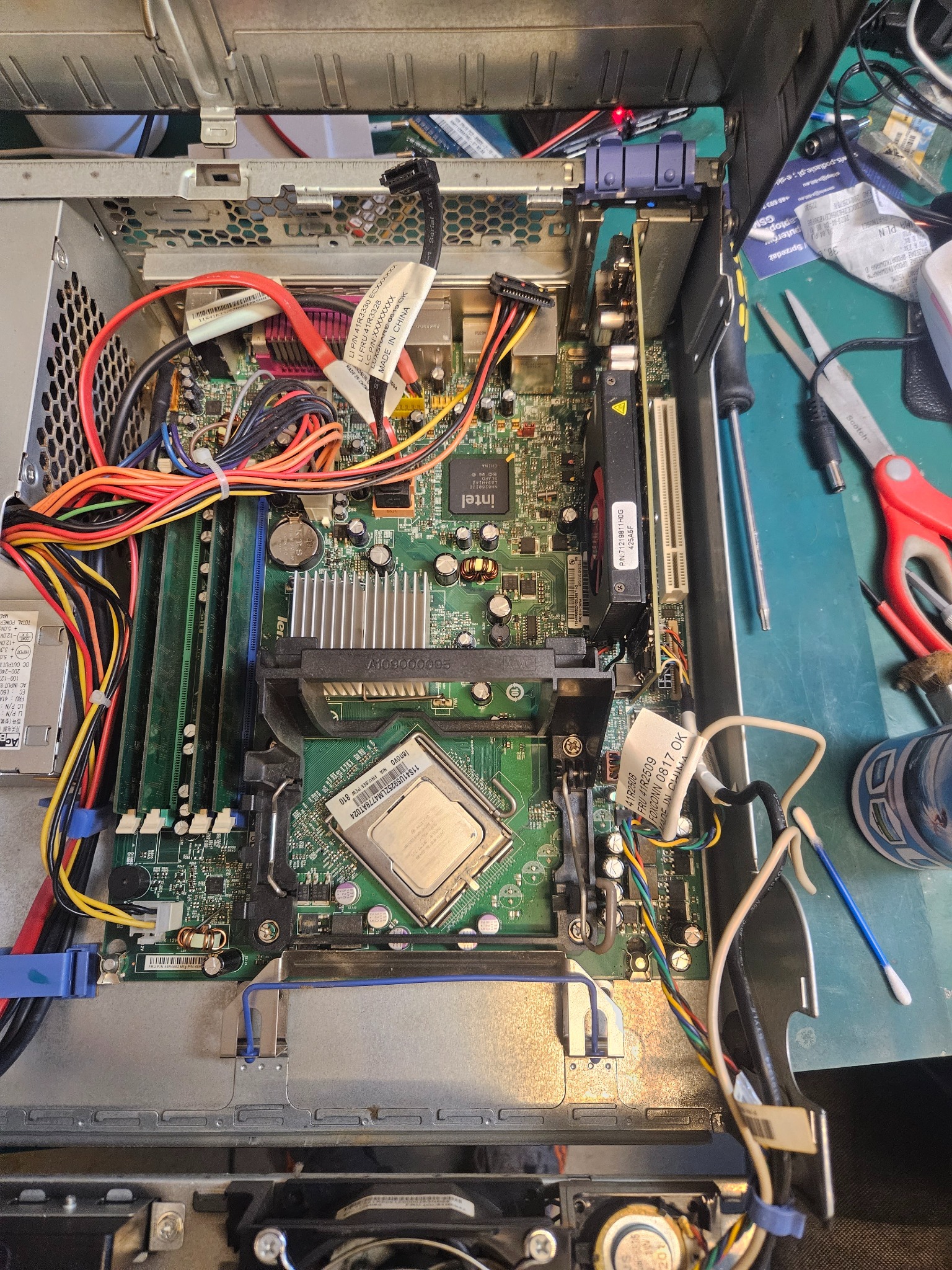 Otwarta obudowa komputera stacjonarnego z widoczną płytą główną, procesorem Intel, pamięcią RAM, kartą graficzną i plątaniną kabli; narzędzia i akcesoria na zielonym tle, sugerujące naprawę...