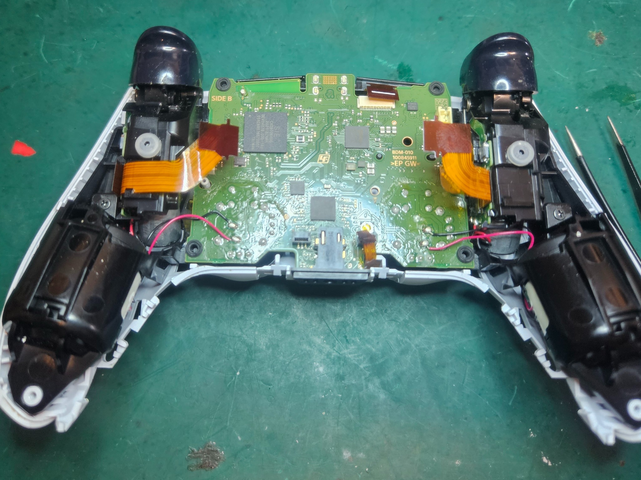 Otwarty kontroler DualSense PlayStation 5 z widoczną płytą główną i elementami wewnętrznymi, leżący na zielonej powierzchni roboczej z narzędziami obok.