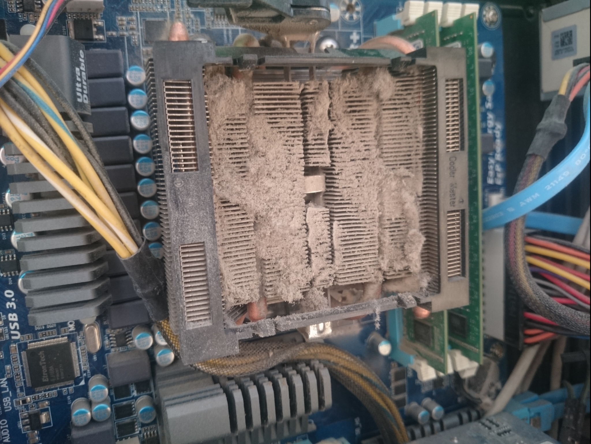 Zabrudzony radiator procesora z widoczną warstwą kurzu na żeberkach, osadzony na płycie głównej komputera stacjonarnego, z widocznymi elementami elektroniki i przewodami.