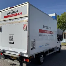 Biała furgonetka z napisem 'Express Car Transport' i kodami QR 'Scan Me' na tylnej i bocznej ścianie, zaparkowana na zewnątrz budynku w słoneczny dzień.