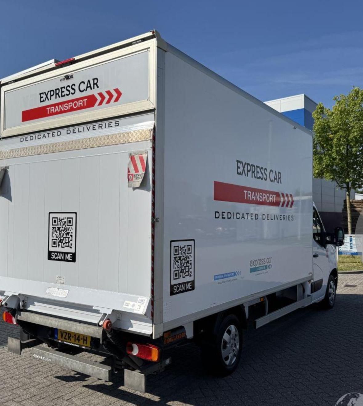 Biała furgonetka z napisem 'Express Car Transport' i kodami QR 'Scan Me' na tylnej i bocznej ścianie, zaparkowana na zewnątrz budynku w słoneczny dzień.