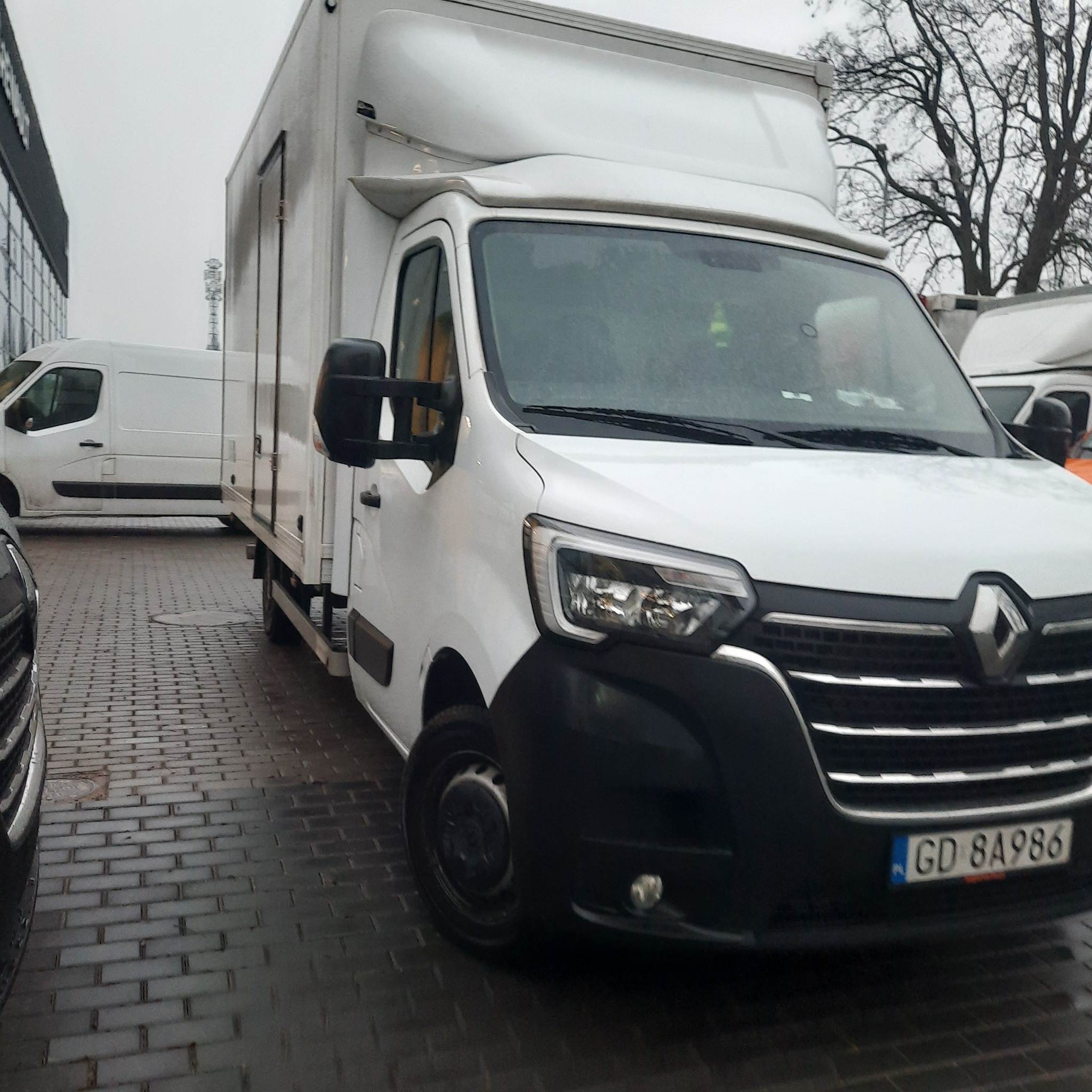 Biały samochód dostawczy Renault Master z wysoką zabudową, zaparkowany na brukowanym parkingu, widoczny numer rejestracyjny GD 8A986, w tle inne samochody dostawcze i budynek.