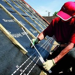 Usługi blacharsko dekarskie Łukasz Maciejowski - Dekarz w czerwonej czapce i zielonych rękawicach mierzy odległość na membranie dachowej Braas Veltite, rozciągniętej na drewnianych łatach, przy pomocy białej miarki.