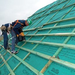 Usługi blacharsko dekarskie Łukasz Maciejowski - Dekarze w uprzężach montują membranę dachową na drewnianej konstrukcji, używając narzędzia pneumatycznego. Widoczne zabezpieczenia, zielony kask i buty sportowe jednego z pracowników. W tle budynek...