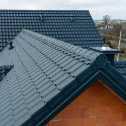 Ciemnoszary, metalowy dach z kominem i wentylacją, widok z g&oacute;ry na tle zabudowań.