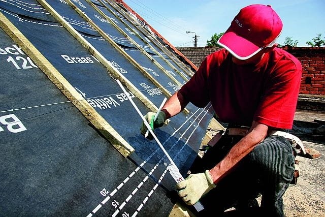 Dekarz w czerwonej czapce i zielonych rękawicach mierzy odległość na membranie dachowej Braas Veltite, rozciągniętej na drewnianych łatach, przy pomocy białej miarki.