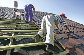 Dekarze pracują na dachu, układając brązową dachówkę na drewnianej konstrukcji. Widoczna czarna membrana dachowa. Jeden z pracowników w czerwonej czapce i rękawicach, drugi w niebieskich spodniach...