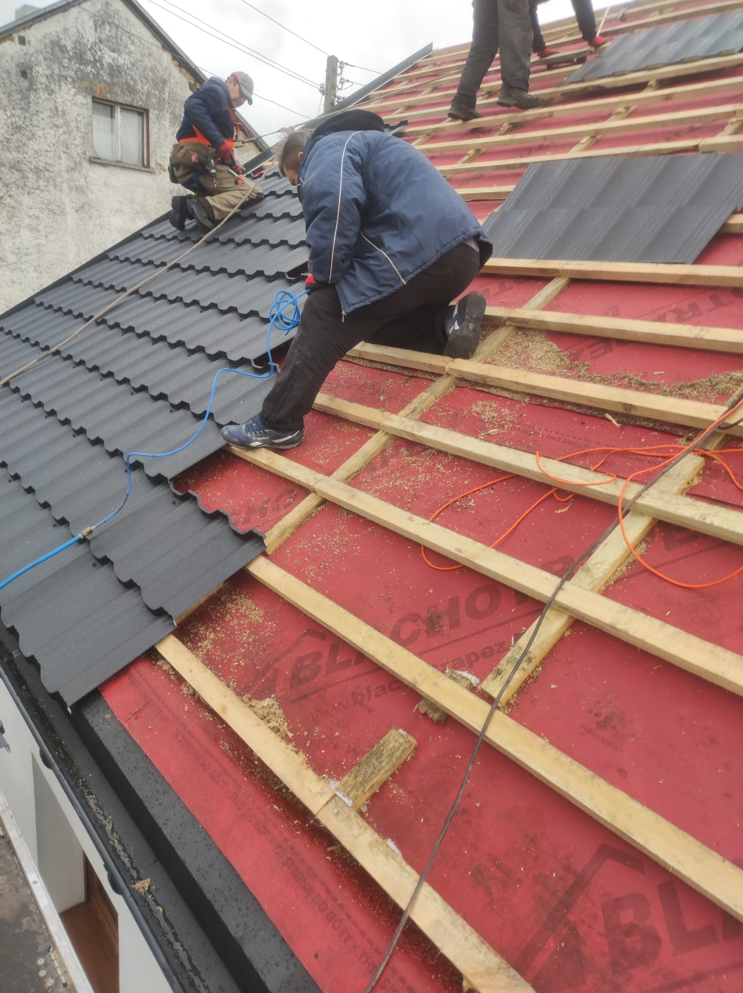 Montaż czarnej blachodachówki na dachu domu jednorodzinnego, widoczne drewniane łaty, czerwona membrana dachowa i pracownicy w trakcie pracy, jeden z nich klęczy na dachu.