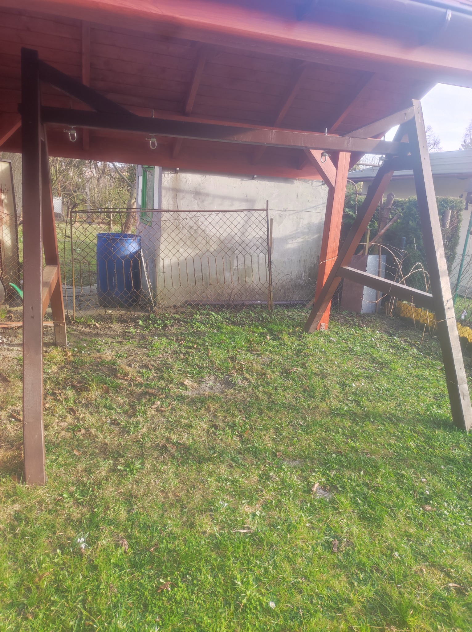 Drewniana konstrukcja zadaszenia w trakcie budowy, widoczne elementy mocujące, w tle niebieska beczka i ogrodzenie z siatki.