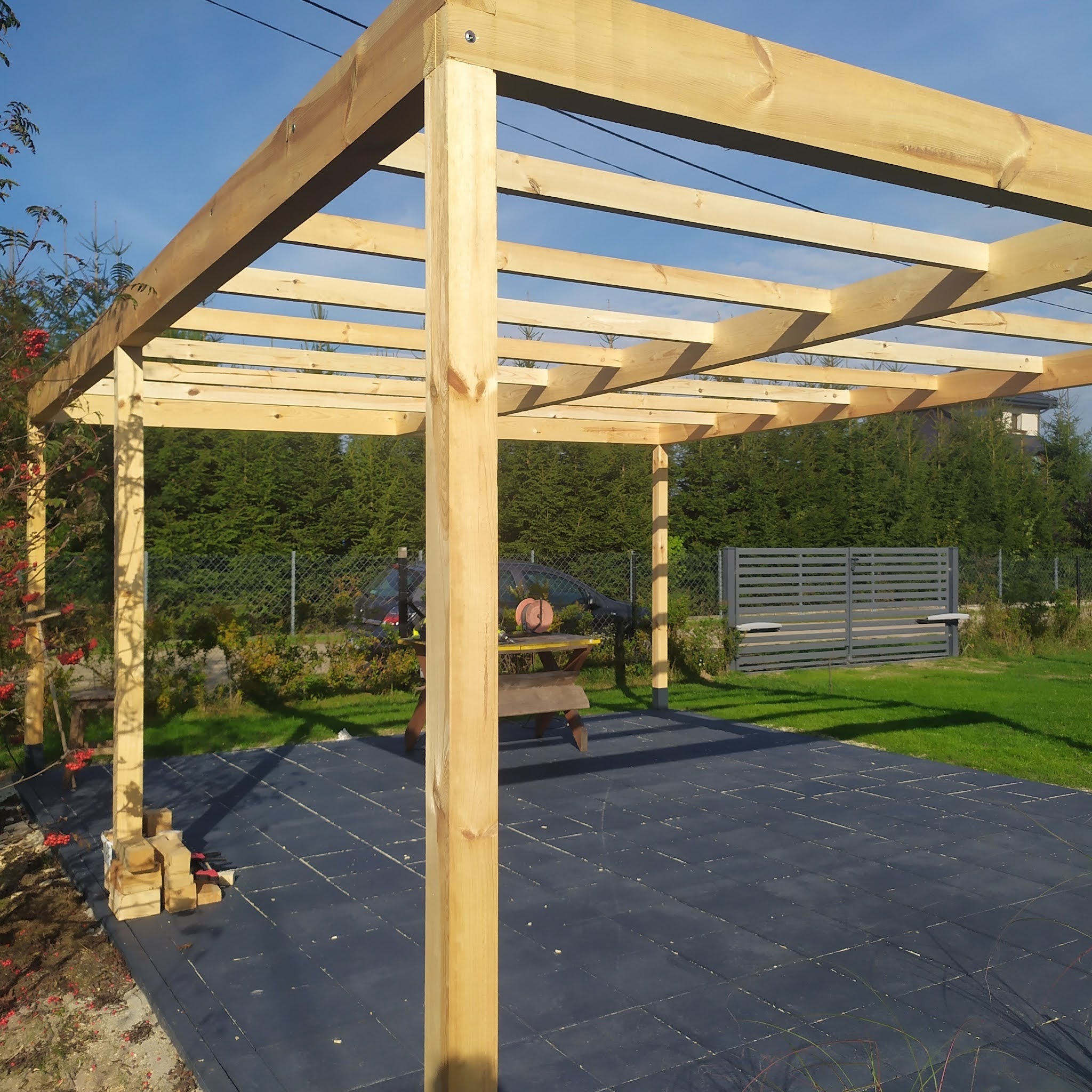 Surowa, drewniana konstrukcja pergoli w trakcie budowy, widoczne nieobrobione belki i słupy, z narzędziami i materiałami budowlanymi w tle, na przygotowanym podłożu z czarnej maty.