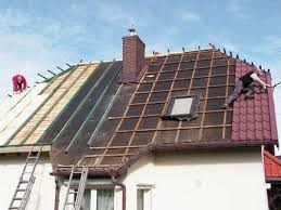 Demontaż starego pokrycia dachowego i montaż nowego pokrycia z blachodachówki w kolorze bordowym. Widoczny fragment dachu z oknem dachowym, rusztowanie oraz dwóch dekarzy pracujących na różnych...