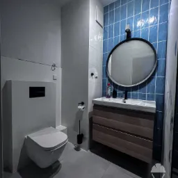 Nowoczesna łazienka po remoncie: wisząca toaleta, umywalka z szafką z ciemnego drewna i okrągłe lustro na ścianie z niebieskich płytek. Minimalistyczny design z szarą podłogą.