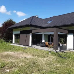 Nowoczesny dom jednorodzinny z białymi ścianami i brązowymi elementami wykończeniowymi, widok z zewnątrz na taras z meblami ogrodowymi, błękitne niebo z chmurami w tle.