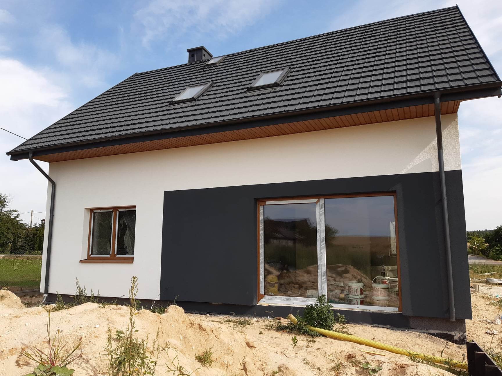 Nowoczesny dom jednorodzinny z dwukolorową elewacją: białą i grafitową, z ciemną dachówką i oknami z brązowymi ramami, widoczne ślady prac wykończeniowych wokół budynku.