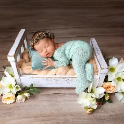 Śpiący noworodek w miętowym kombinezonie i wianku na głowie, leżący w małym, białym łóżeczku na miękkim kocyku, z dekoracją z białych i brzoskwiniowych kwiatów.