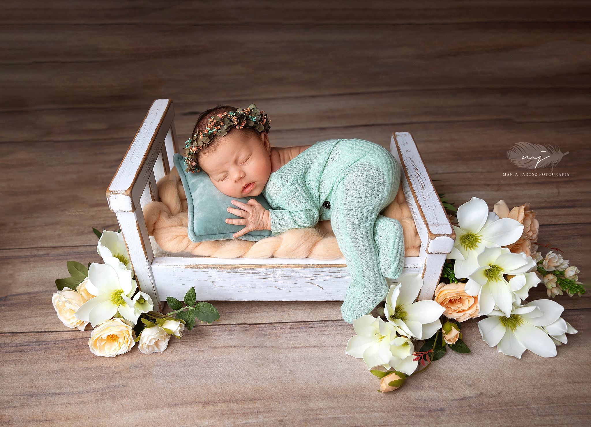 Śpiący noworodek w miętowym kombinezonie i wianku na głowie, leżący w małym, białym łóżeczku na miękkim kocyku, z dekoracją z białych i brzoskwiniowych kwiatów.