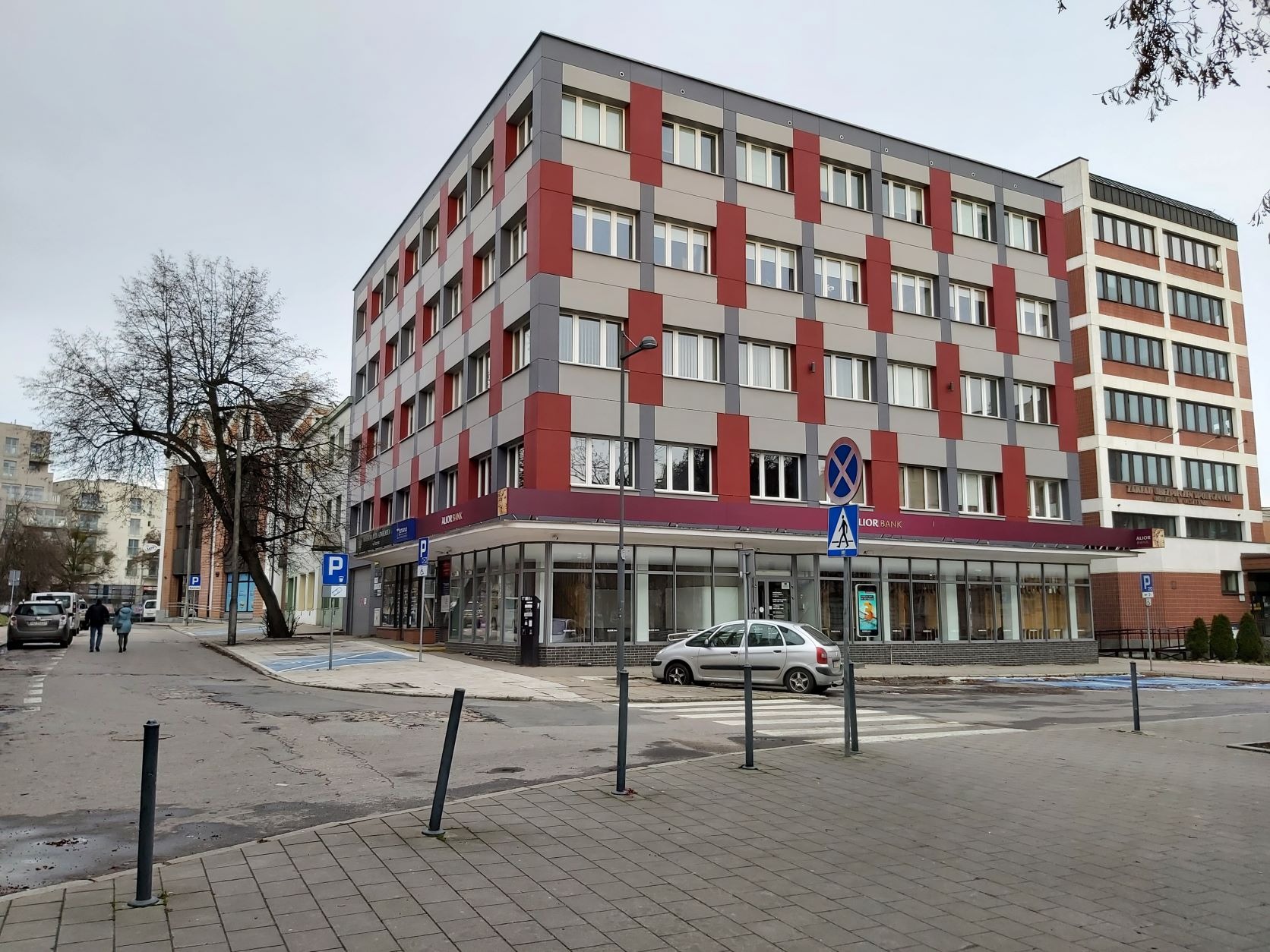 Nowoczesny budynek biurowy z elewacją w szaro-czerwone pasy, widoczny z poziomu ulicy z przejściem dla pieszych i znakami drogowymi.