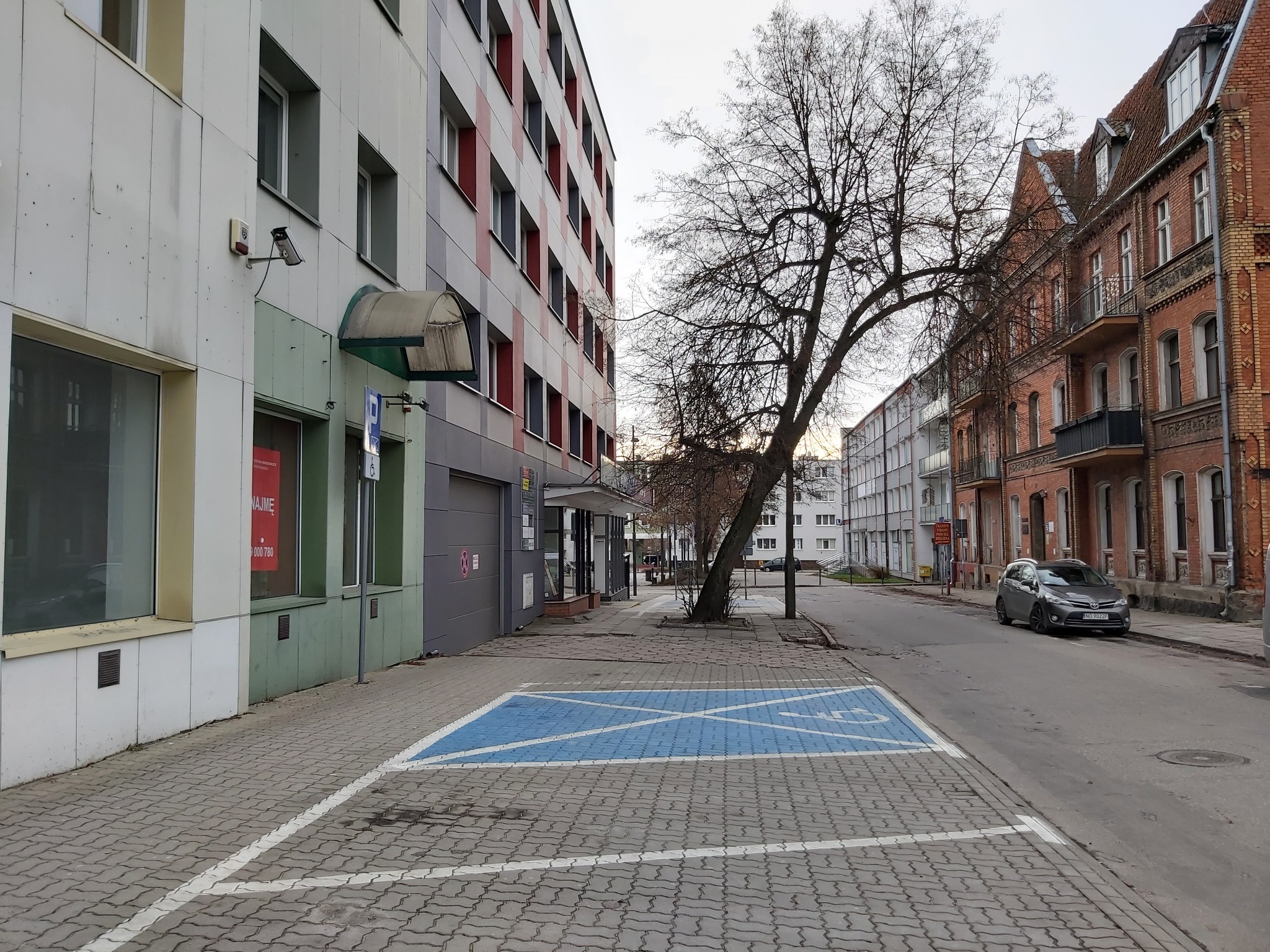 Ulica miejska z budynkami o różnej architekturze, w tym nowoczesny biurowiec i kamienica z czerwonej cegły, niebieskie miejsce parkingowe dla niepełnosprawnych na pierwszym planie.