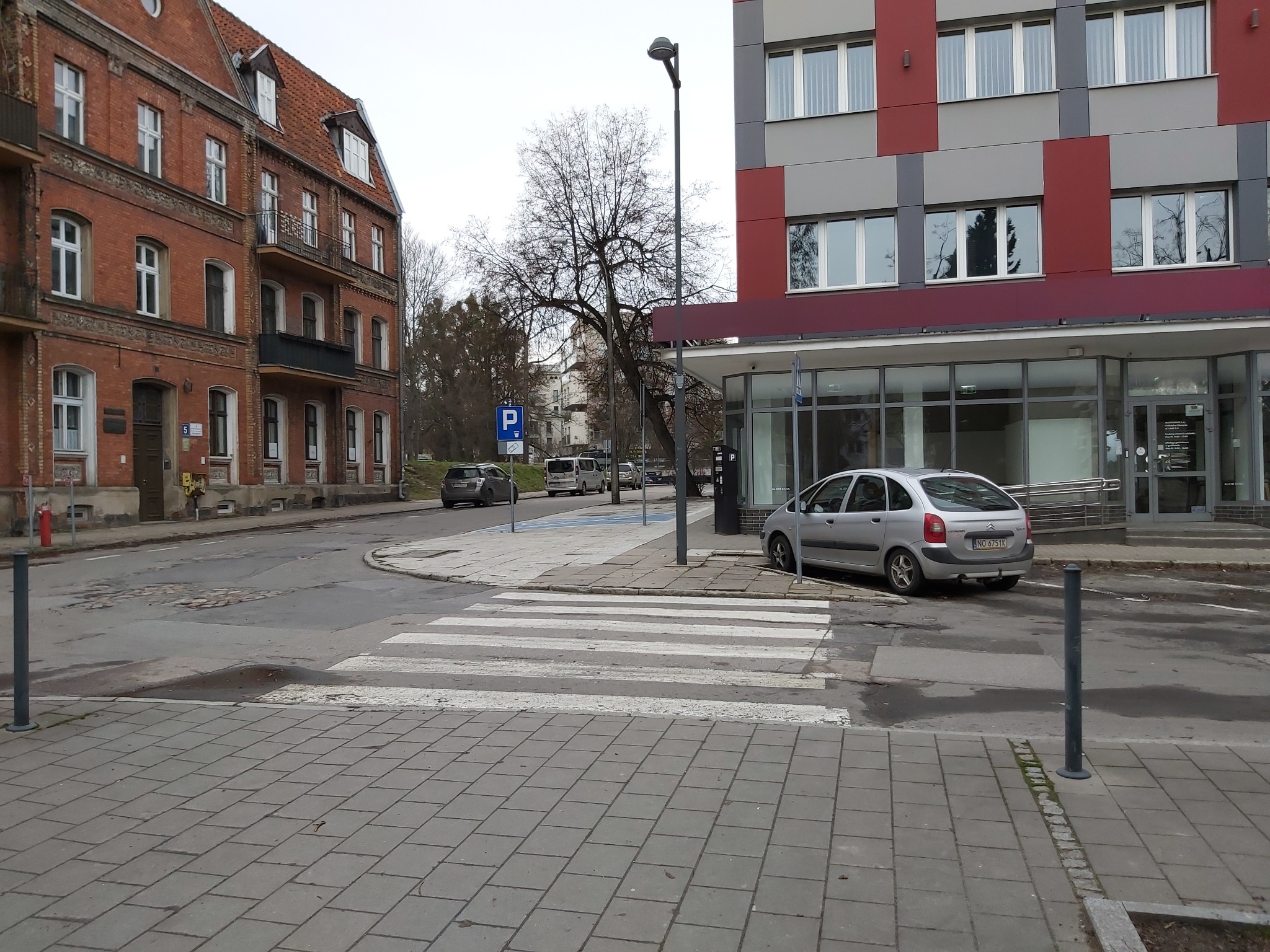 Ulica z przejściem dla pieszych, zaparkowanym samochodem i budynkami w tle, w tym budynek z szaro-czerwonymi panelami i przeszklonym parterem, oraz starsza kamienica z czerwonej cegły.