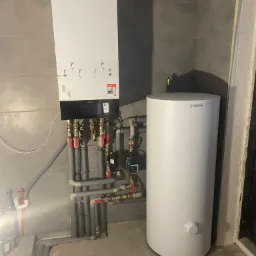 Kocioł grzewczy Viessmann wiszący na szarej ścianie z widocznymi rurami i zaworami, obok biały zasobnik wody. Surowy stan wykończenia pomieszczenia.