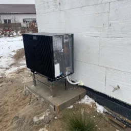 Czarna pompa ciepła Viessmann na metalowym stelażu, zamontowana przy ocieplonej ścianie budynku w trakcie instalacji, widoczne białe rury i pianka uszczelniająca, w tle częściowo ośnieżony teren.