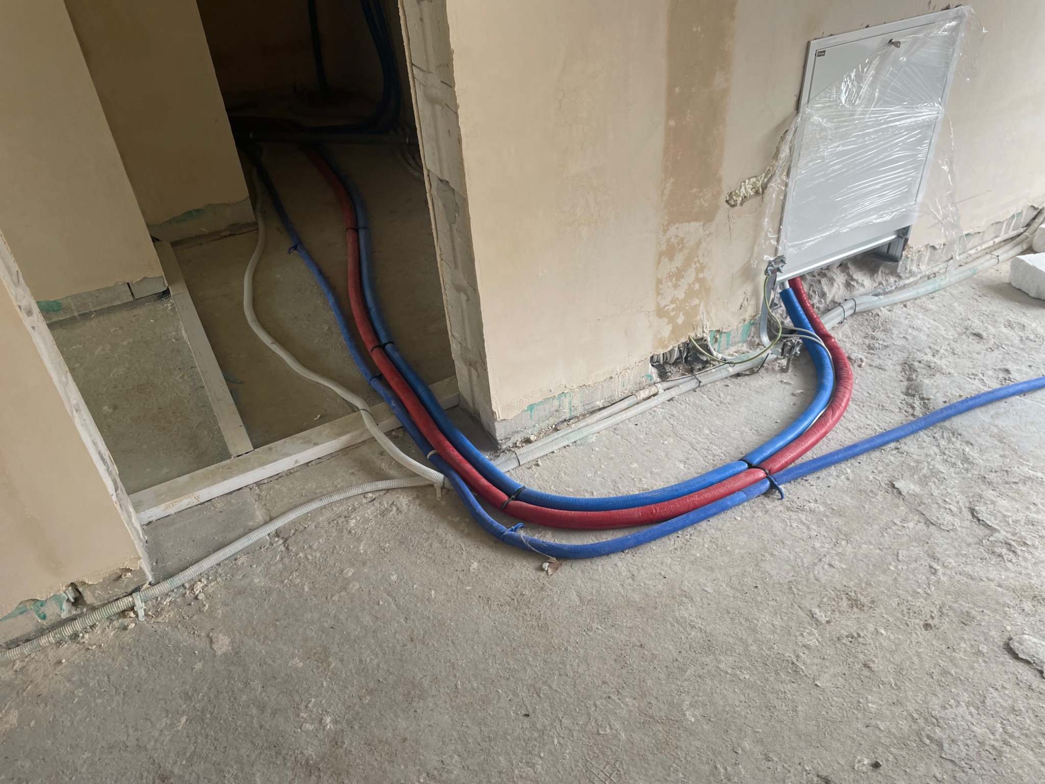 Wiązka rur instalacyjnych w kolorach niebieskim i czerwonym, wychodząca z otworu w ścianie w surowym stanie, prowadząca po betonowej podłodze. Na ścianie widoczna skrzynka instalacyjna...