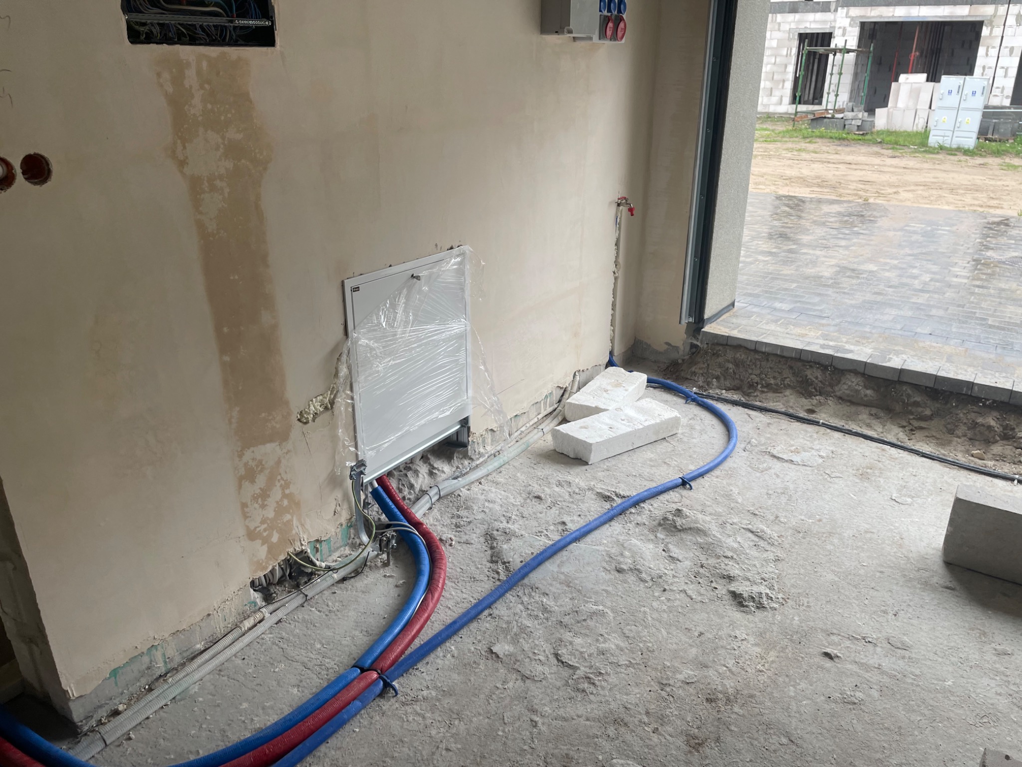 Wnętrze w trakcie prac instalacyjnych: widoczne rury w izolacji czerwono-niebieskiej prowadzone wzdłuż ściany, skrzynka rozdzielcza zabezpieczona folią, wystające przewody elektryczne...