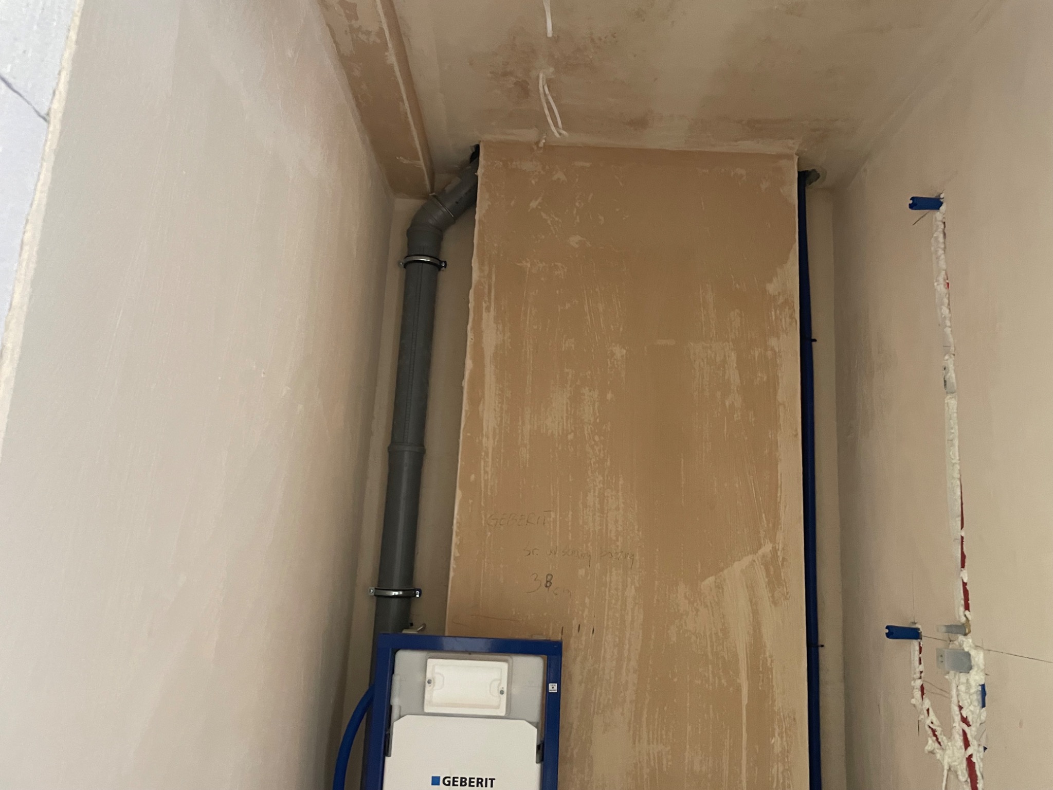 Montaż stelaża podtynkowego Geberit z szarą rurą odpływową w surowym wnętrzu łazienki, widoczne ślady prac budowlanych na ścianach i suficie.