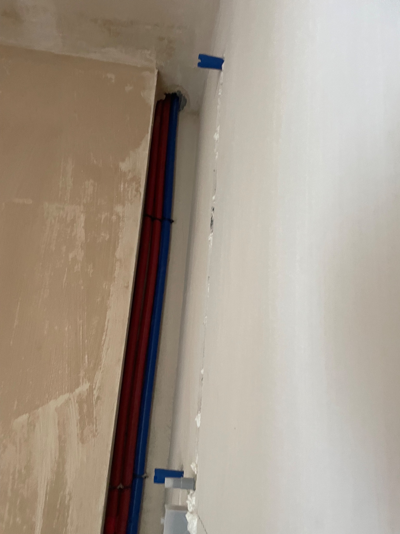 Wnęka w ścianie z pionowo ułożonymi rurami w kolorach czerwonym i niebieskim, zabezpieczonymi niebieską taśmą malarską, ściany w stanie surowym przed wykończeniem.