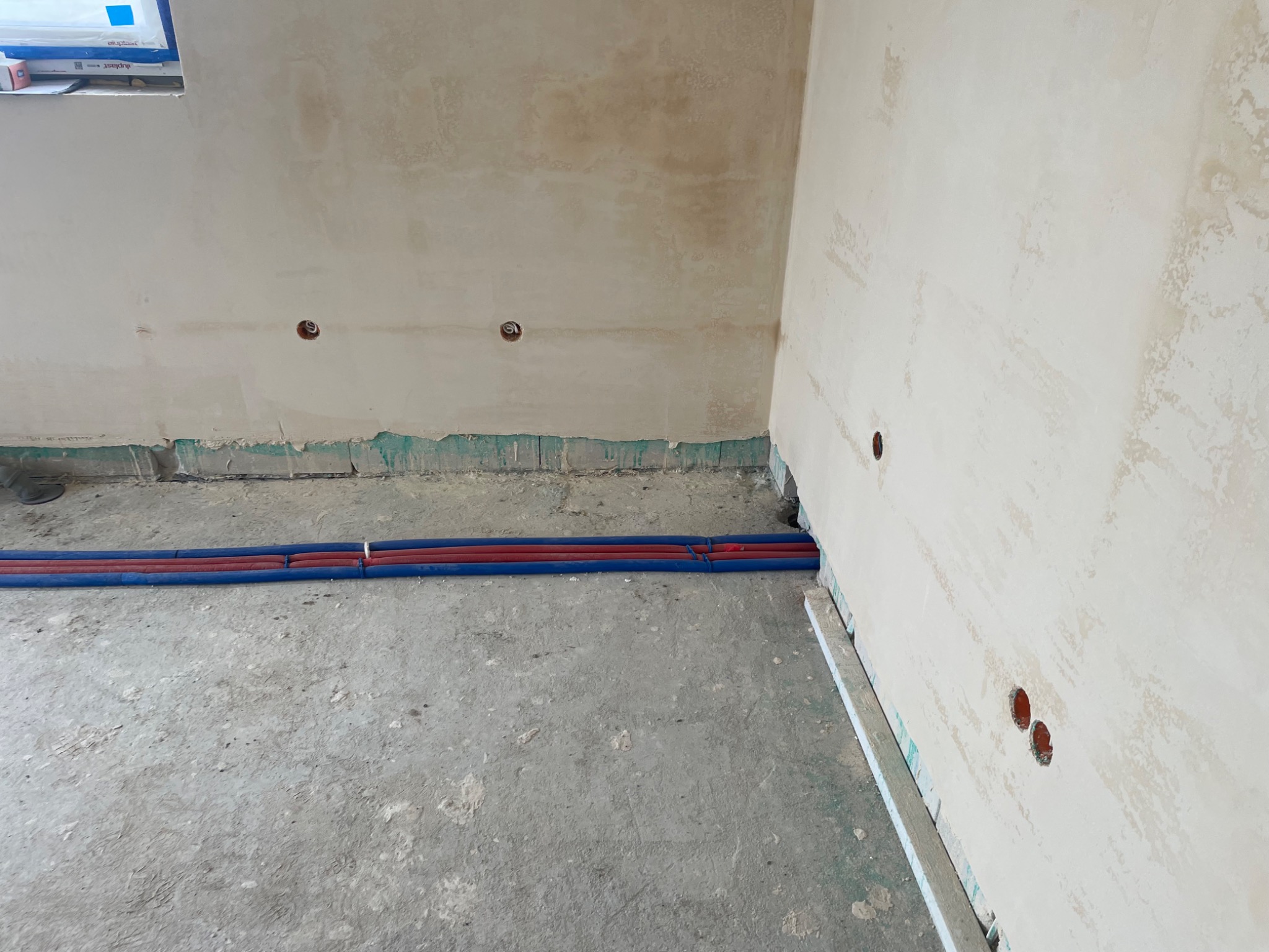 Rury instalacji wodnej w izolacji, ułożone wzdłuż ściany w surowym stanie, widoczne puszki elektryczne w ścianie.