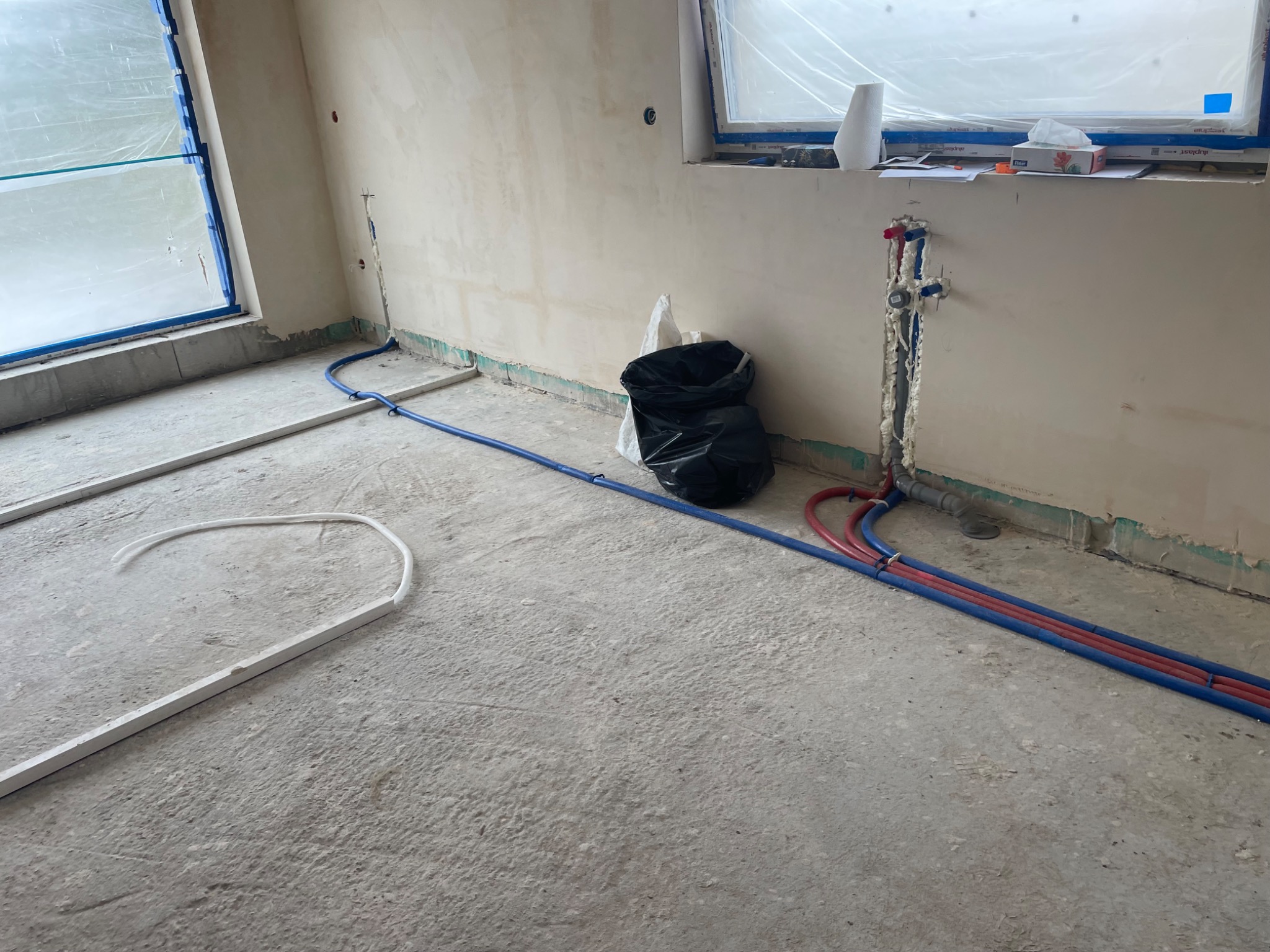Surowy stan wnętrza w trakcie prac instalacyjnych: widoczne rury hydrauliczne (czerwone i niebieskie) prowadzone po betonowej podłodze, białe listwy przypodłogowe, okna zabezpieczone folią, ściany...